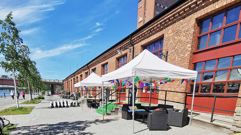 Hamnmagasinet utifrån en solig sommardag. Utanför entrén finns soffgrupper och paviljoner.